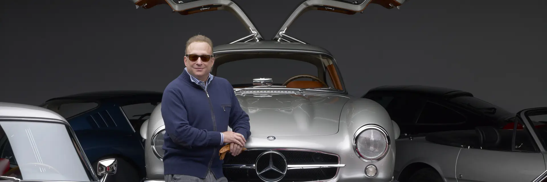 Miles Nadal tall white older man in his 50s60s wearing classic Ray Ban plasticframed sunglasses a blue zipup sweater with a striped buttonup collared shirt leaning against the front hood of his silver MercedesBenz 300 SL Gullwing with the gullwing doors opened a part of the front drivers side of a silver Aston Martin and the front driver side profile of a red car.