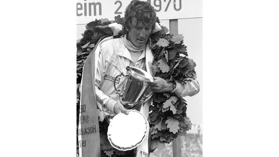 Jochen Rindt stands on the podium holding his final 1st-place trophy at the 1970 German Grand Prix. This victory would secure his World Championship later that year.