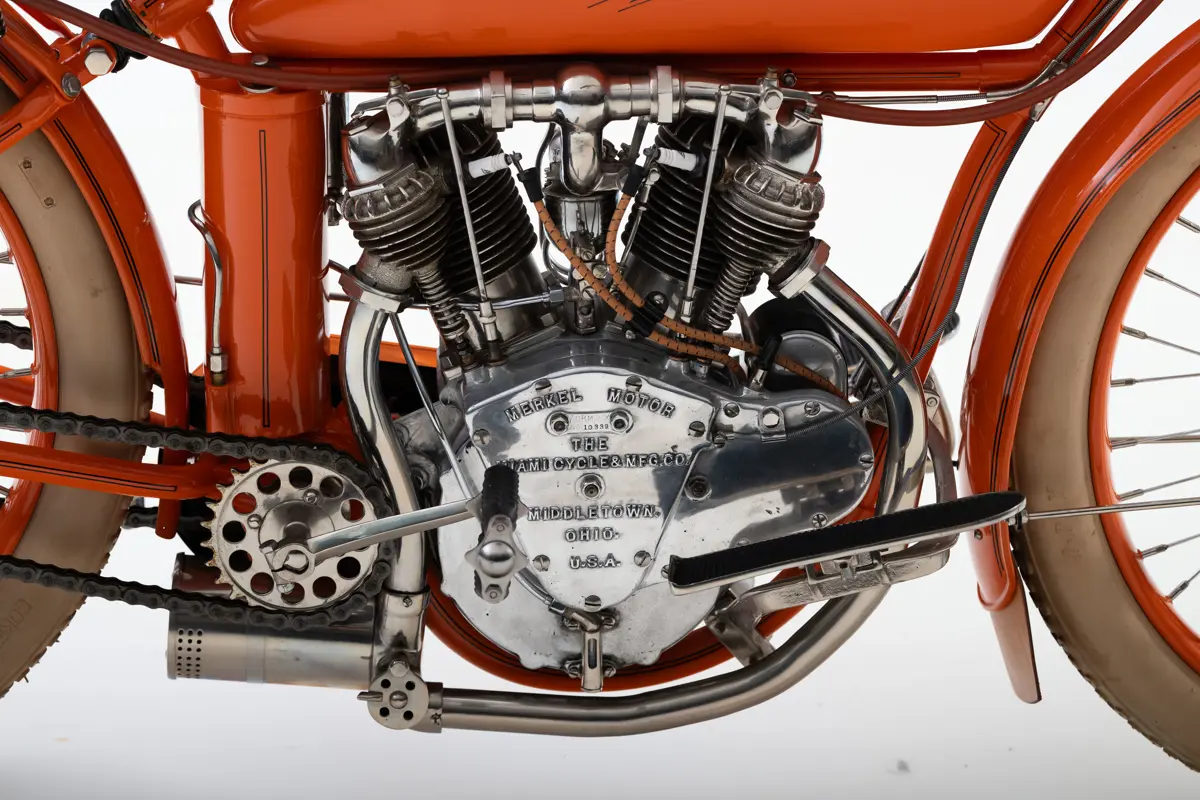 A close up of the V-twin engine with two distinctive cylinder chambers spotted with air cooling metal fins surrounding them. An upside down bell-shaped stamp is plaited to the engine reading ‘Merkel Motor. The Miami Cycle &amp;amp; MFG. CO. Middletown Ohio USA