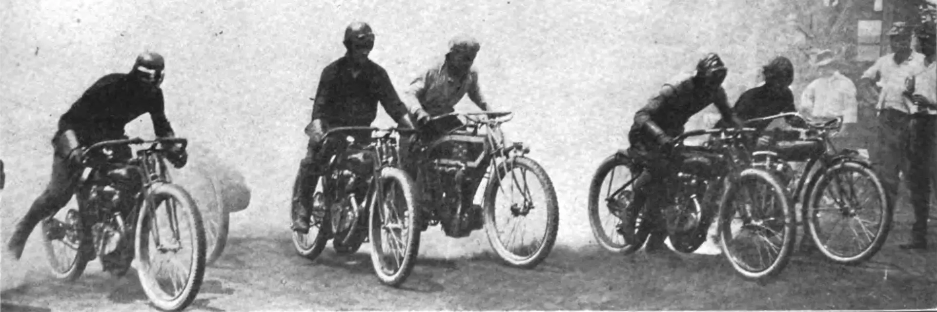 1913 image taken of the Flying Merkel Racing Team in black grey and white. You can clearly see five riders on their motorcycles lined up across a dirt track with a cloud of smoke behind emitted from the bikes behind them. You can just make out some faint silhouettes of a crowd sitting to the left of the riders or the right side and rear of the image