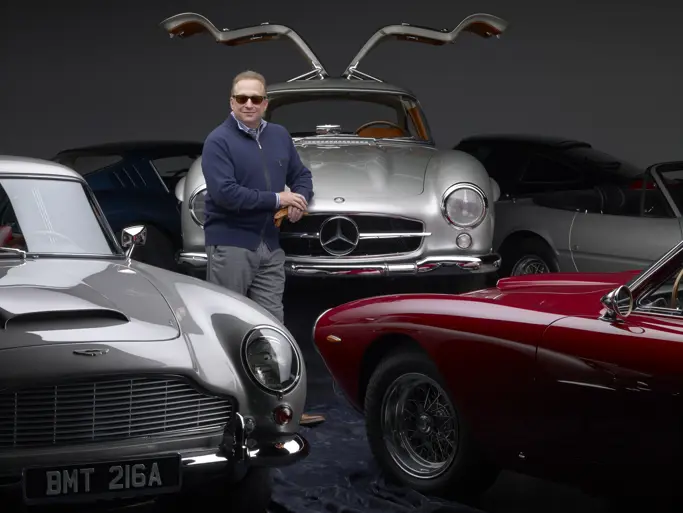 Miles Nadal tall white older man in his 50s60s wearing classic Ray Ban plasticframed sunglasses a blue zipup sweater with a striped buttonup collared shirt leaning against the front hood of his silver MercedesBenz 300 SL Gullwing with the gullwing doors opened a part of the front drivers side of a silver Aston Martin and the front driver side profile of a red car.