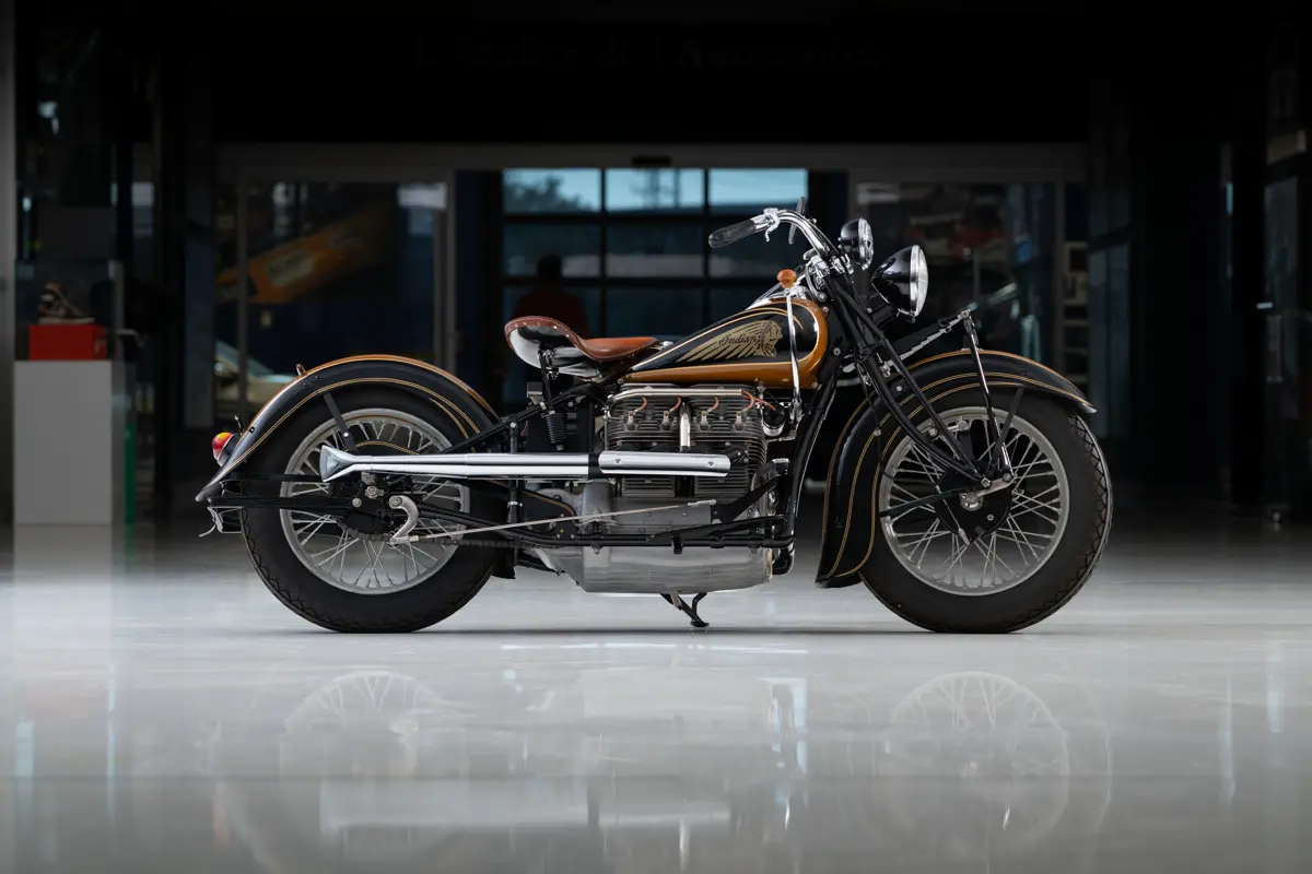 A left-side profile of an older, 1938 Indian Four motorcycle model with a longer wheelbase, chrome exhaust, and gold accents on the trim. The engine is an air-cooled inline four with orange-gold plug wires. A gold Indian chief is painted on the tank. Up front is a small circular light off of the handle bar, with another, larger circular headlight. It’s on a side stand on a white ceramic flooring in a dimly lit garage with camera strobe lighting