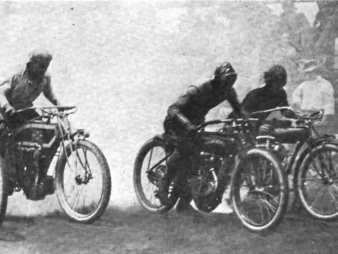1913 image taken of the Flying Merkel Racing Team in black grey and white. You can clearly see five riders on their motorcycles lined up across a dirt track with a cloud of smoke behind emitted from the bikes behind them. You can just make out some faint silhouettes of a crowd sitting to the left of the riders or the right side and rear of the image