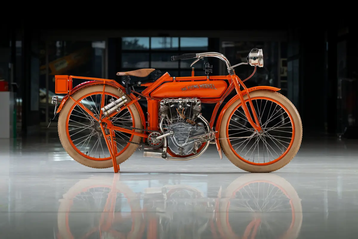 An older motorcycle that looks like a hefty early 20th century bicycle with an engine fitted to it. The 1914 Flying Merkel Model 471 wearing a slightly red-orange paint, with an all cap font on the gas tank reading ‘The Flying Merkel.’ the tires are white rubber fitted to spoked wheels with orange rims. The bicycle theme is apparent with the chain-driven, bicycle pedals, set just behind the small v-twin motor