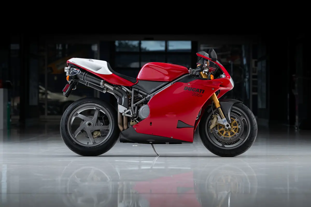 A right-side profile of a 2001 Ducati 996R from the Dare to Dream Collection, taken in a dimmed garage with strobe lighting. The Ducati looks racy with most components raked forward like the racing seat and rider position. It wears a red carbon fiber faring extending from the front fairing over the headlight down to below the engine, with ‘Ducati 996-R’ marked. Peaking out from under the tail which is wearing white carbon fiber, is a set of Termignoni exhausts